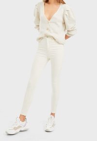 Cárdigan blanco de punto con mangas abullonadas y cierre de botones, combinado con jeans skinny blancos y zapatillas gruesas con acentos en beige y gris.