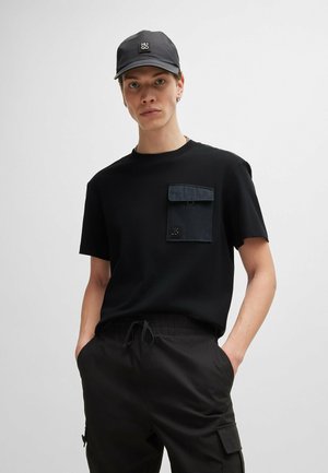 Camiseta negra con un bolsillo en el pecho que cuenta con una solapa y un logo, combinada con pantalones cargo negros. El tejido parece suave y ligero.