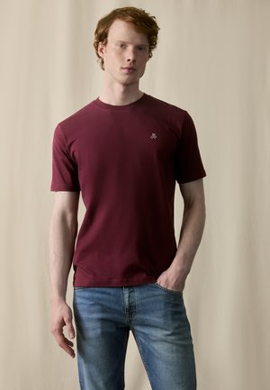 Burgunderfarbenes T-Shirt mit kurzen Ärmeln und Rundhalsausschnitt, mit kleinem Logo auf der linken Brust, kombiniert mit hellblauen Jeans. Glatte Stofftextur.