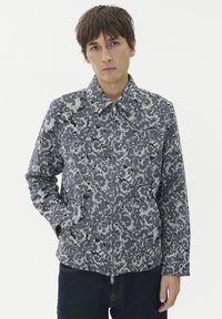 Mlad moški s kratkimi rjavimi lasmi, oblečen v zadrgo z okrasnim cvetličnim vzorcem v sivi in črni barvi ter temno modre kavbojke, na bež ozadju.