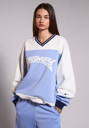 Sudadera de color azul claro con acentos blancos, diseño de cuello en v y un gráfico destacado de "Primed". Presenta detalles de canalé en el cuello y los puños de color marino.