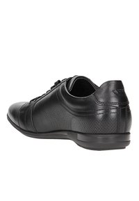 Černá kožená bota s kulatou špičkou, texturovanými perforovanými detaily, hladkým povrchem a nízkým gumovým podešvem. Snímatelné tkaničky jsou součástí.