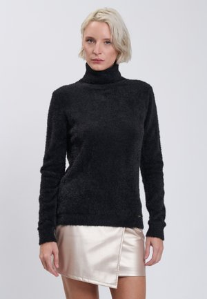 Donna con capelli biondi corti che indossa un maglione nero peloso a collo alto e una minigonna beige asimmetrica lucida su uno sfondo semplice.
