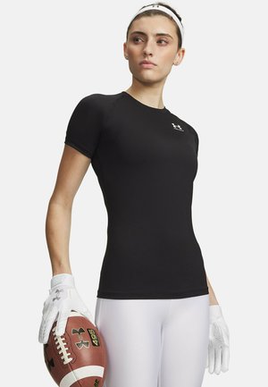 Atletische vrouw in zwart T-shirt met korte mouwen en witte leggings houdt een voetbal vast met witte handschoenen, kijkend naar de zijkant tegen een effen achtergrond.