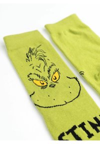 Calze verdi con il volto di un personaggio in stile cartoon, caratterizzato da occhi gialli, contorni neri e la scritta "STINK" sotto il design. Texture del tessuto morbida.