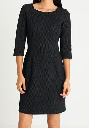 Zwart, geribbeld knielang jurkje met driekwart mouwen en een vierkante halslijn, gedragen door een staande persoon.