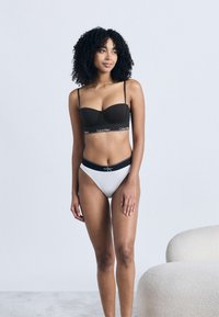 Γυναίκα με σγουρά μαλλιά, φοράει μαύρο σουτιέν Calvin Klein και λευκό σλιπ ψηλής κοπής, στέκεται κοντά σε μια κρεμ πολυθρόνα με υφή.