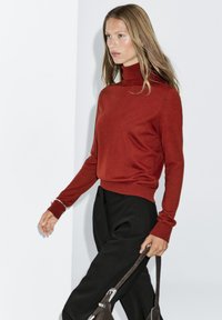 Sweter z golfem w odcieniu rdzy, z dopasowanymi mankietami, zestawiony z luźnymi czarnymi spodniami. Miękka tekstura, elegancki krój i minimalistyczny design.