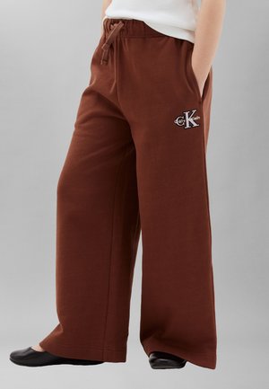 Person som har på seg løse brune joggebukser med Calvin Klein-logo på venstre lår, kombinert med svarte slip-on sko og hvit topp.