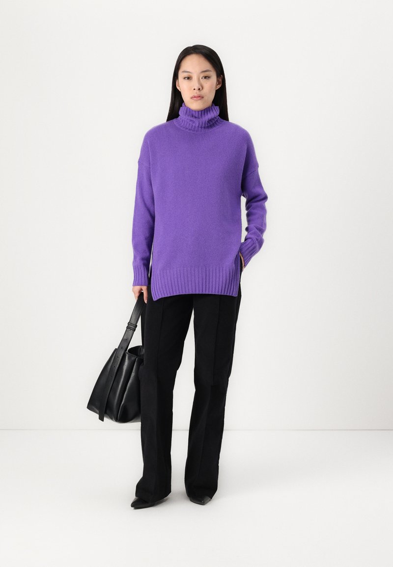 Lila Rollkragenpullover mit geripptem Saum, kombiniert mit schwarzen geraden Hosen. Das Model hält eine schwarze Handtasche. Einfaches Design, weiche Textur.