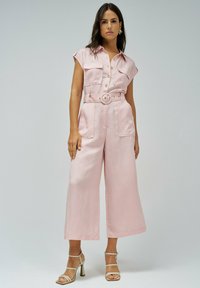 Mujer de pie vestida con un mono sin mangas de color rosa claro, ceñido a la cintura y con pantalones anchos, y sandalias de tacón alto beige, contra un fondo liso.