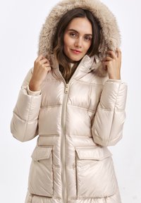 Woman adjusting hood on beige puffer jacket with fur trim, smiling softly, against a plain white background.