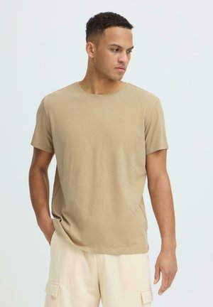 Junger Mann mit kurzem lockigem Haar, der ein beiges Kurzarm-T-Shirt und cremefarbene Cargo-Shorts trägt, schaut zur Seite und hat eine Hand in der Tasche.