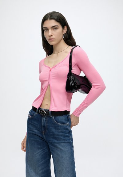 Personne aux longs cheveux foncés portant un haut rose à manches longues, un jean bleu, une ceinture noire et une petite sacoche noire en bandoulière, debout sur fond blanc.
