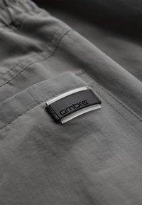 Primer plano de un bolsillo de tela gris con una etiqueta rectangular negra y transparente con la palabra «ombre» cosida en el borde.