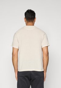Camiseta beige de manga corta con cuello redondo y corte casual, confeccionada en una mezcla de algodón texturizado, mostrada desde la parte de atrás.
