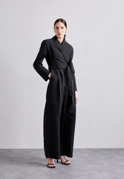 Salopetă neagră, ajustată, cu mâneci lungi, design înfășurat, curea asortată, picioare largi și textură netedă. Asortată cu tocuri negre cu bretele.