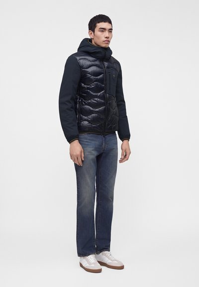 Jeune homme debout portant une veste matelassée noire avec capuche, un jean bleu et des baskets blanches sur un fond blanc uni.