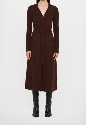 Nainen pukeutuneena pitkähihainen, tummanruskea neulemekko, jossa on napit edessä, yhdistettynä mustiin polven korkeisiin saappaisiin, seisoo valkoista taustaa vasten.