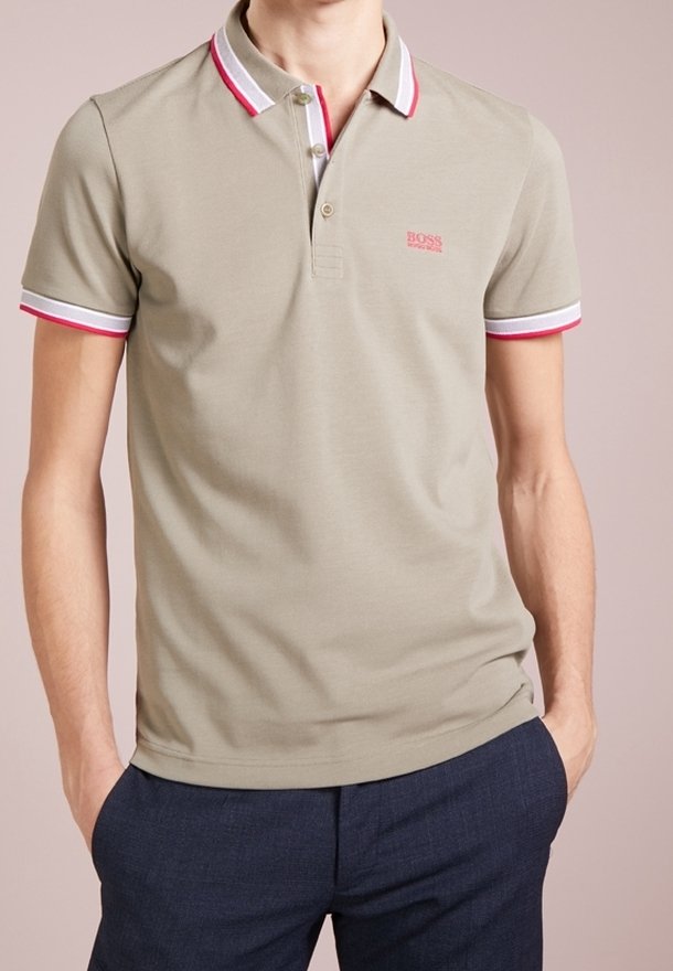 Homme portant un polo beige à manches courtes avec un col et des bords de manches rayés blanc et rouge, associé à un pantalon bleu foncé.
