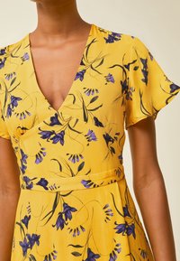 Żółta sukienka z dekoltem w serek, krótkimi falbanastymi rękawami i fioletowymi kwiatowymi wzorami. Posiada ściągniętą talię i miękką, gładką teksturę materiału.