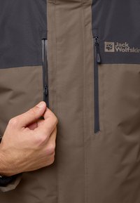 Tweekleurige jas in donkergrijs en lichtbruin, met ritssluitingen en een logo. De stof lijkt glad en weerbestendig.