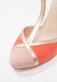 Koralový a světle růžový semišový vysoký podpatek s špičatou špičkou, zdobený zlatými akcenty na překřížených páscích a hladkou texturou.