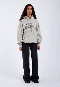 Light gray hooded sweatshirt in soft fabric with a front pocket, featuring black cursive logo on the chest, paired with black wide-leg jeans.