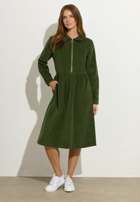 Groene corduroy jurk met een kraag, ritssluiting aan de voorkant en een gathered taille. Lange mouwen en zijzakken, gecombineerd met witte sneakers.