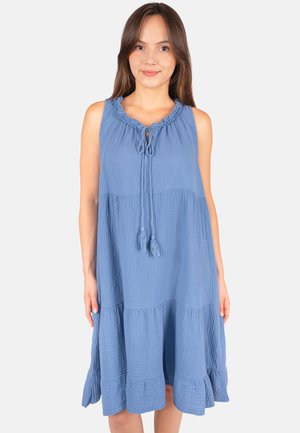 Femme portant une robe bleue sans manches longueur genou avec un col volanté et des liens à pompons à l'avant, debout sur un fond clair.