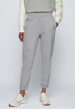Person iført lysegrå joggingbukser, hvide sneakers og en langærmet top, stående med den ene hånd i lommen.