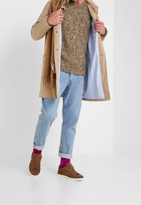 Gabardina beige sobre un suéter de punto multicolor, jeans de mezclilla azul claros y zapatillas marrones. Los accesorios incluyen una bufanda color beige y calcetas magentas.