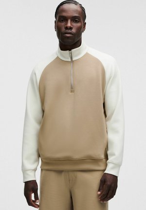 Hombre con pelo trenzado que lleva un jersey beige y blanco de manga larga con cuello con media cremallera y pantalones beige a juego.
