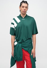 Jaunas asmuo su pintais plaukais, dėvintis žalią Adidas marškinėlį su baltomis juostelėmis ir raudonus kelnes, žalia striukė surišta aplink liemenį.