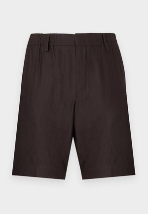 Homme debout, les mains dans les poches, portant un t-shirt bleu clair, un short marron foncé, des chaussettes blanches et des baskets en daim beige sur fond blanc.