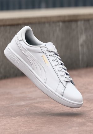 Zapatilla blanca de Puma con cordones, mostrando el perfil lateral y el logo, flotando sobre una superficie exterior lisa con un fondo difuminado de piedras.