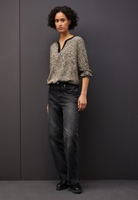 Bluzka w cętki z lamparta w odcieniach beżu i czerni, zestawiona z luźnymi czarnymi dżinsami. Modelka stoi na tle ciemnoszarym, nosząc czarne buty.