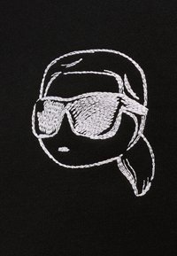 Weiß gesticktes stilisiertes Gesicht mit nach hinten gekämmtem Haar, Sonnenbrille und einem Pferdeschwanz auf schwarzem Stoffhintergrund.