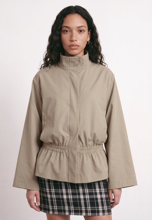 Une femme présente une veste beige à col montant et à la taille froncée, assortie d’une jupe à carreaux noirs et blancs, sur un fond uni.