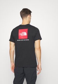 Czarny bawełniany t-shirt z czerwonym kwadratowym nadrukiem, na którym znajduje się logo "THE NORTH FACE" nad napisem "NEVER STOP EXPLORING" na plecach.