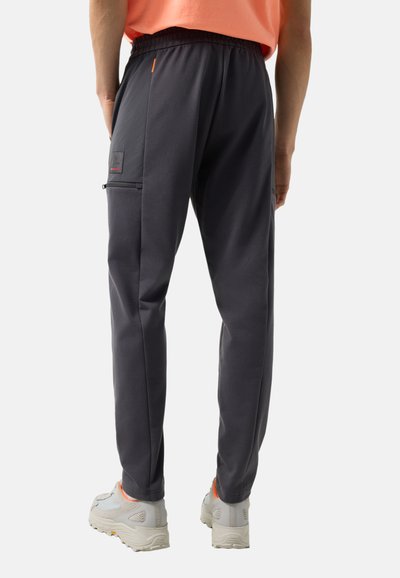 Osoba ubrana w czarne, zwężane spodnie dresowe z suwakiem po boku oraz białe sneakersy z pomarańczowymi akcentami, widziana od tyłu.