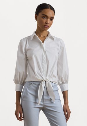 Chemise blanche à manches ballon avec un devant noué, au toucher lisse. Associée à un pantalon tailor bleu clair.