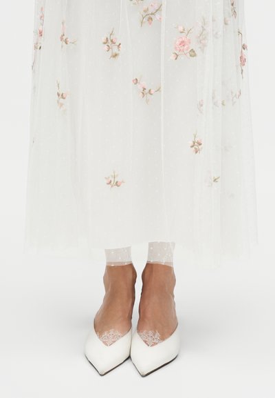 Pieds portant des chaussettes blanches transparentes brodées de fleurs et des chaussures blanches à bout pointu sous une robe blanche avec une broderie de fleurs roses.