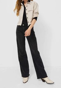 Beige corduroy jas met knopen, zwarte aansluitende top, hoge zwarte jeans en witte hakken met golvende details.