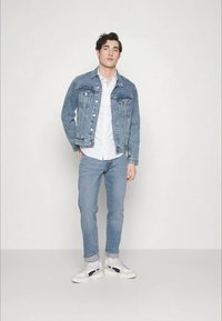 Corta-vento de ganga com um deslavado claro, camisa branca por baixo, combinado com jeans azul claro e sapatilhas brancas com detalhes em azul-marinho.