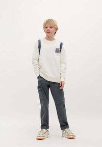 Felpa bianca con una grafica sul petto, abbinata a pantaloni cargo grigi con tasche laterali e risvolti, indossata con sneaker.