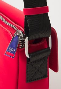 Red synthetic bag featuring a silver zipper with a blue tag, black strap, and textured black accents near the attachment points.