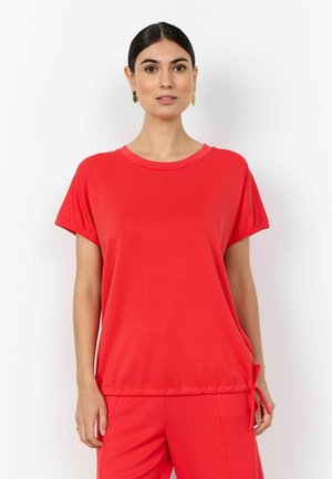 Femeie cu păr închis la culoare, purtând cercei aurii, un top roșu cu mâneci scurte și legătură laterală și pantaloni roșii asortați, stând în fața unui fundal alb.