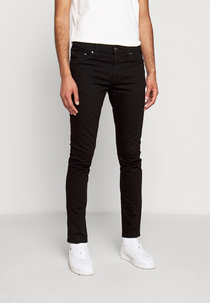 Black slim-fit jeans made of denim, featuring a classic five-pocket design and metal hardware. Worn with white sneakers and a light shirt.