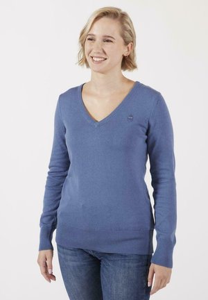 Pull à col V bleu clair, avec des poignets et un ourlet côtelés, texture de tricot douce, arborant un petit logo sur la poitrine gauche. Porté avec un jean foncé.